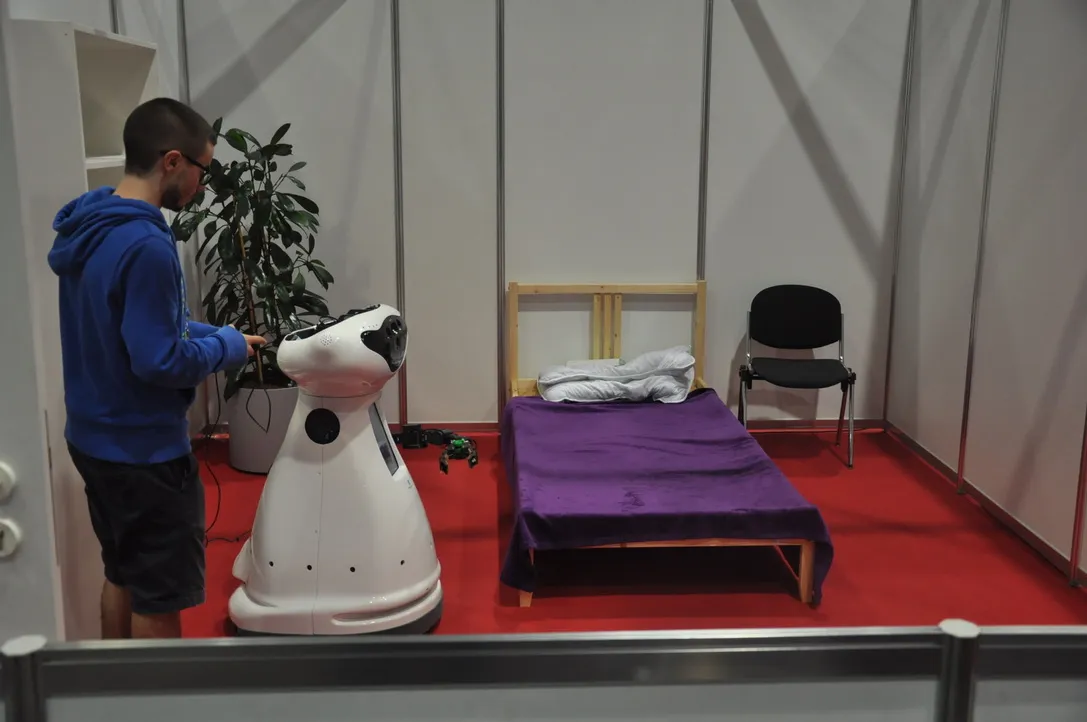 Nuno in a blue hoodie working on a white service robot in a mock bedroom at a RoboCup arena with a red floor.
